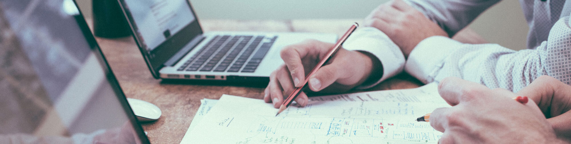 Arme zweier Personen mit Stiften am Schreibtisch, zwischen Laptops mit handschriftlichen Papieren befasst